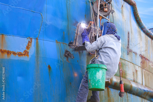 Welder rappelling with ladder to welding metal hull surface of the old fishing boat during maintenance and improvement work in shipyard area