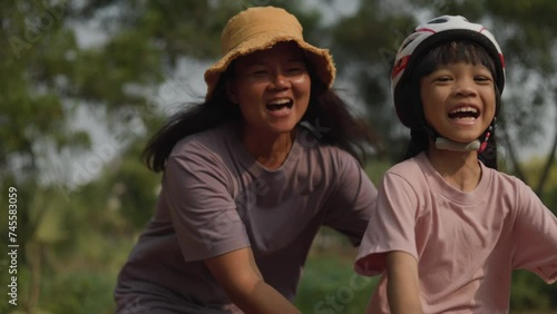 A handheld video of an asian mother take care her duaghter while she rides a red bike in the park with happy and smile on both of their face in the weekend in green woods background.