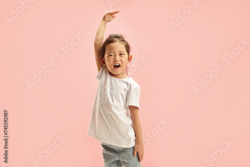 Cheerful Asian ethnicity little girl playfully stretches arm upwards, showing her height, denoting how she grew up, reaching for the sky, symbolizing growth, ambition, standing on pink isolated wall photo