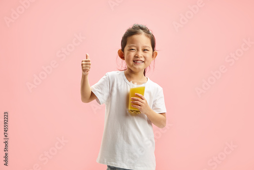 Happy Smiling Little Brunette Child Girl Drinking Orange Juice From Glass, Expresses Pleasure Positively Showing Thumb up Standing On Pink Isolated photo
