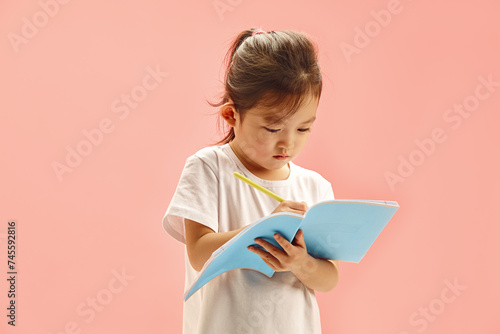 Japanese apperance child concentrating on writes to copybook, pen in hand, against pink background. photo