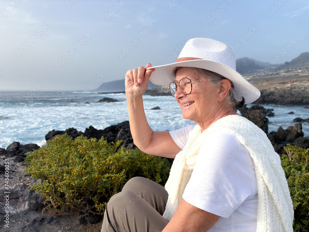 Side view of senior smiling woman sitting on a rocky beach watching ocean waves crashing with white foam. Concept of relaxed elderly person admiring the power of nature
