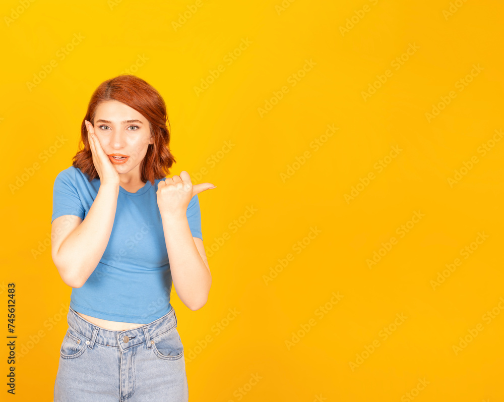 Shocked young caucasian red bob hair woman pointing aside copy space with thumb. Touching her cheek with hand, open mouth. Yellow studio background. Demonstrating workspace area. Shocked young caucasian red bob hair woman pointing aside copy space with thumb. Touching her cheek with hand, open mouth. Yellow studio background. Demonstrating workspace area.