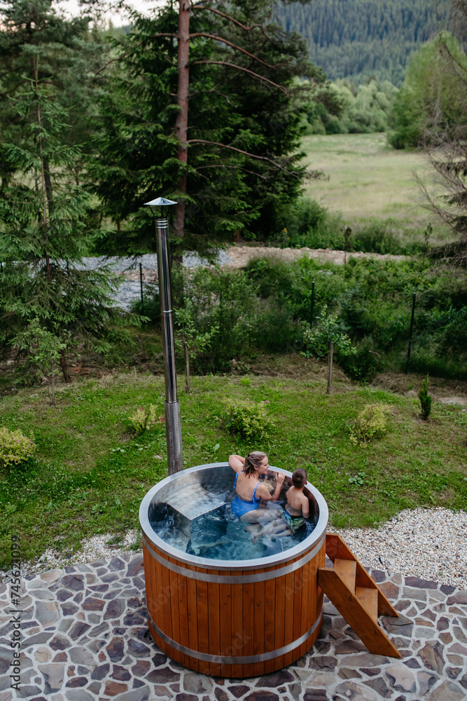 Mother with her little son enjoying bathing in wooden barrel hot tub in the terrace of the cottage. Wooden bathtub with a fireplace to burn wood and heat water.
