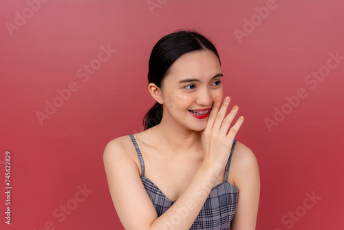 A young Asian woman whispers with a smile, hand on mouth, signifying secrecy or gossip against a plain background.