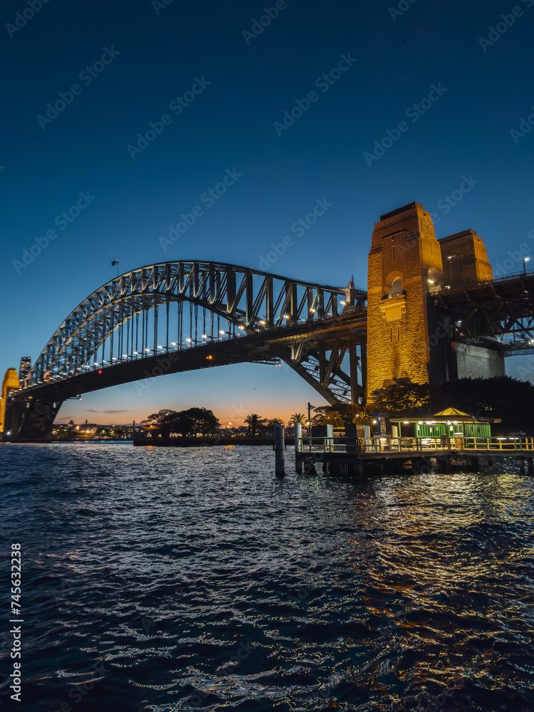 Obraz premium Sydney by night and at sunset is a beautiful sight to behold. The city skyline, complete with the iconic Harbour Bridge, is a picture-perfect view of Australia.
