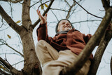 Happy boy playing on tree during first spring days. Kid climbing tree, sitting on branch and smiling. Outdoor, garden activity for young children.
