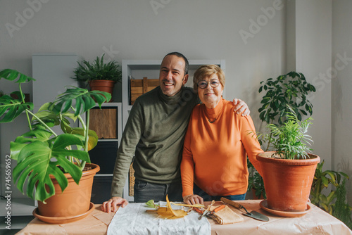 Smiling son with arm around mother standing near plants on table at home photo