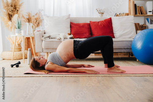 Pregnant woman lying on exercise mat practicing bridge pose in living room photo