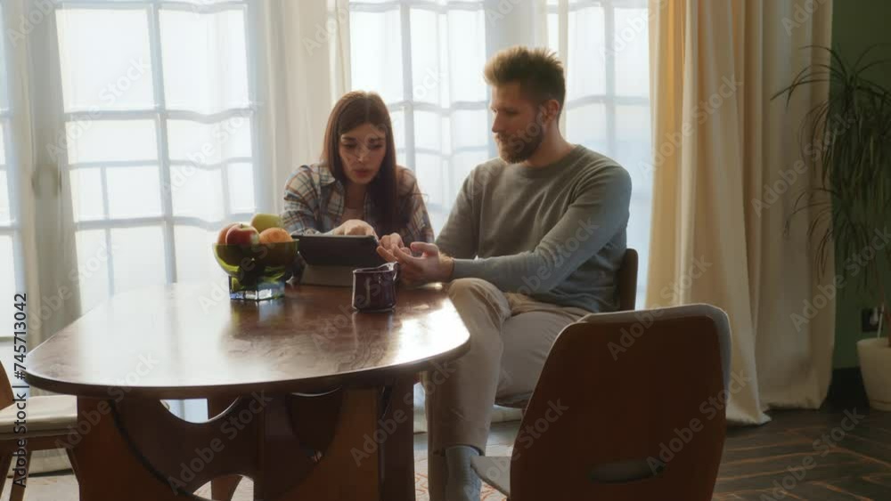 young love couple sit at table watching to tablet screen together, chatting at home at sunny morning. romantic lovers relax in living room, using app on pad, smiling and laughing. young love couple sit at table watching to tablet screen together, chatting at home at sunny morning. romantic lovers relax in living room, using app on pad, smiling and laughing.