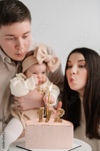 Family blows out candle on cake. Father, mother hold hands little kid celebration six month old baby girl closeup. Dad and Mom hug cute baby isolated on white wall. Creative birthday party, having fun