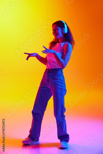 Positive young lady dancing while listening music in headphones against gradient studio background in mixed neon light. Concept of human emotions, youth culture, self-expression, technology. Ad