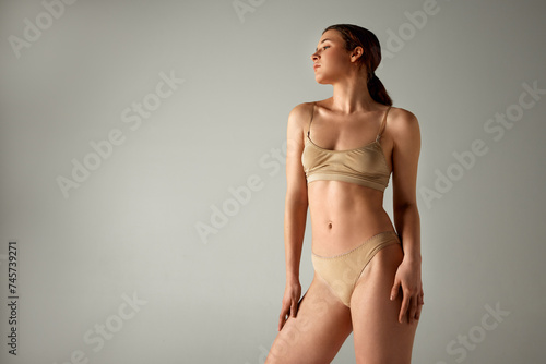 Beautiful young woman with ponytail, slim, fit body posing in underwear against grey studio background. Self-love and care. Concept of body and health care, female beauty, wellness