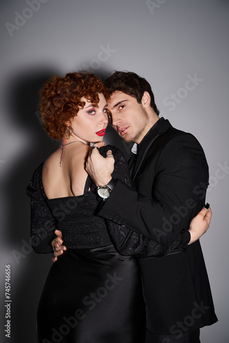 good looking passionate couple in black chic attires hugging and looking at camera on gray backdrop