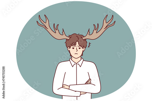 Disgruntled guy with deer horns on head stands with arms crossed and looks at screen with displeasure. Metaphor of man who suffered from infidelity of girlfriend or adultery wife