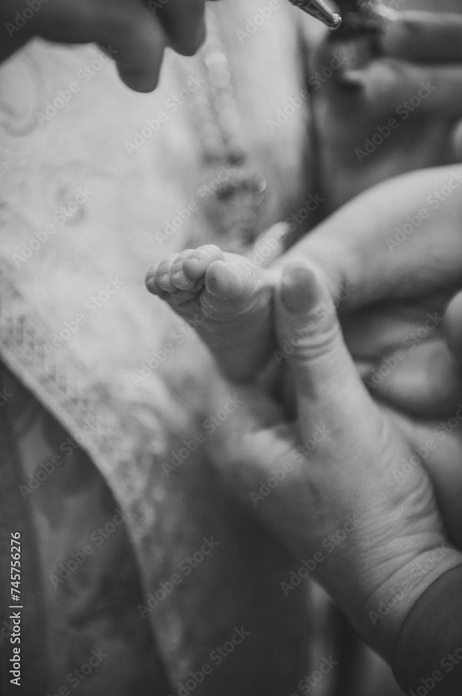 Black and white photo anointing. Unction at baptism. Priest and godfather stretch hands to legs. Temple, Orthodoxy. Closeup of tiny baby feet, the sacrament of baptism ceremony. Godmother holds child.