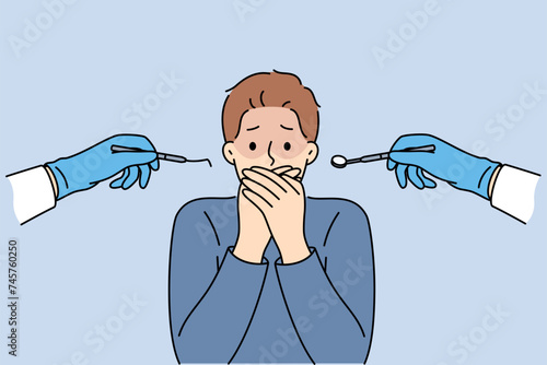 Man suffering from dentophobia closes mouth and stands near hands with dental devices. Guy is afraid of doctors at dental clinic, and feels panic, not allowing caries to be treated