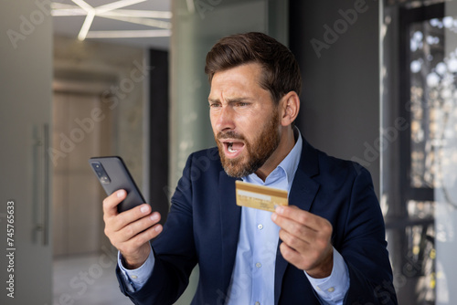 Close-up photo of angry young businessman in suit standing in office, holding golden credit card in hand and looking disappointedly at mobile phone screen