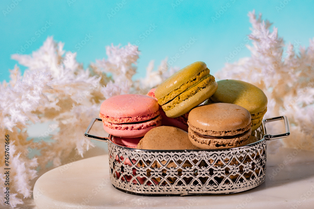 Colorful macaroons arranged in a silver bowl on a stone marble table among boho-style flowers and on a blue background Colorful macaroons arranged in a silver bowl on a stone marble table among boho-style flowers and on a blue background