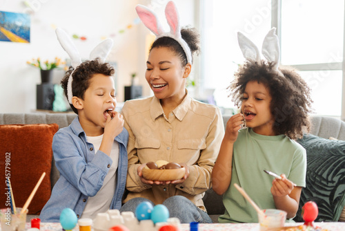 Portrait happy latin family eating chocolate eggs at home. Smiling mother and kids wearing bunny ears celebration Easter together. Holiday activity concept photo