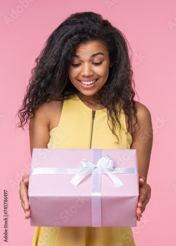 Studio shot of happy smiling beautiful birthday girl looking at the decorated present box in her hands with excited face expression, isolatd over pastel pink background