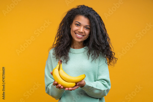 Studio shot of happy smiling attractive young woman posing with a bunch of fresh ripe bananas in hands isolated over bright colored orange yellow background