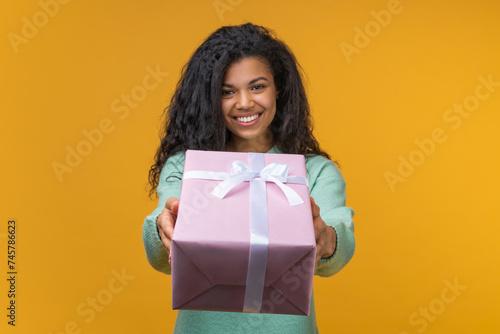 Studio shot of attractive smiling young woman reaching the present box to the camera, isolated over bright colored orange yellow background