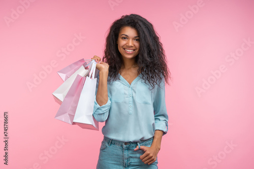 Studio portrait of young stylish smiling confident young woman holding a bunch of shoppers on hand and posing over pastel pink background