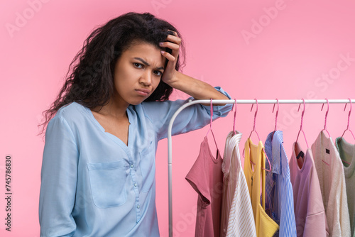 Half-length portrait of dissapointed young woman standing in showroom near the rail of pastel colored clothing unable to find fitting look