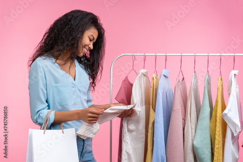 Studio shot of happy smiling stylish elegant young woman choosing outfit hanging on rack in clothing store, isolated over pastel pink background