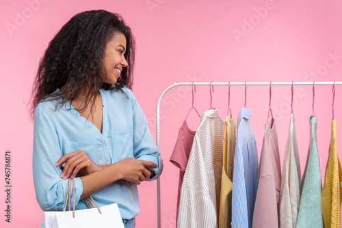 Studio portrait of attractive stylish smiling elegant young woman with shopping bags is choosing apparel hanging on rack  in clothing store isolated over pastel pink background