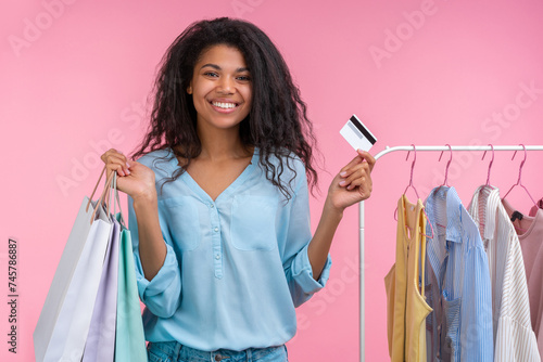Studio portrait of joyful attractive stylish elegant young woman posing with with shopping bags and credit card in hands while standing near the rack of pastel colored clothes, isolated on pink backgr