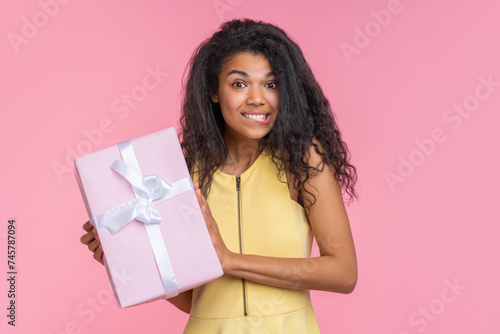 Studio portrait of cute coquette girl posing over pastel pink background with decorated gift box in hands