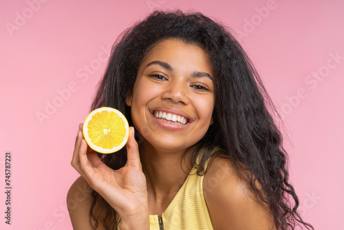 Close up studio portrait of beautiful happy young woman with charming smile posing over pastel pink background with a lemon cut in a half in hand