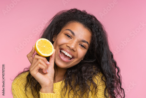 Close up studio portrait of cute happy smiling girl posing over pastel pink background with a lemon cut in a half in hand
