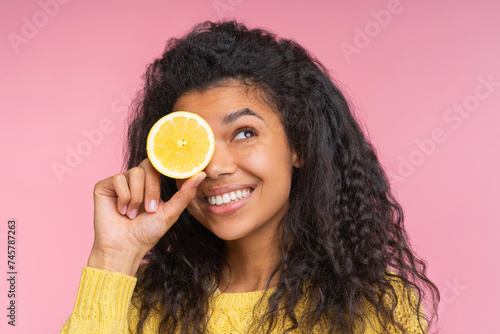 Close up studio portrait of attractive coquette young woman with charming smile posing over pastel pink background with a lemon cut in a half in hand