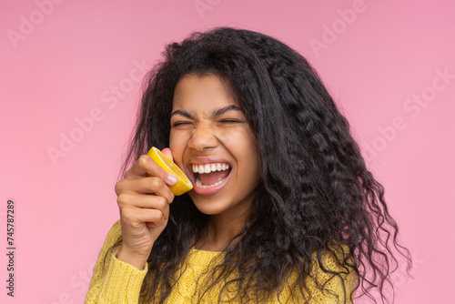 Portrait of beautiful young african american woman posing over pastel pink background pretending to bite a lemon cut in a half