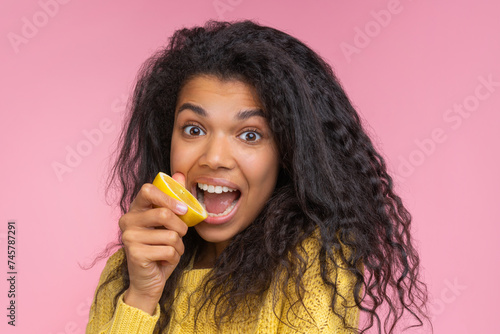 Close up portrait of cute and funny african american girl posing over pastel pink background pretending to bite a lemon cut in a half