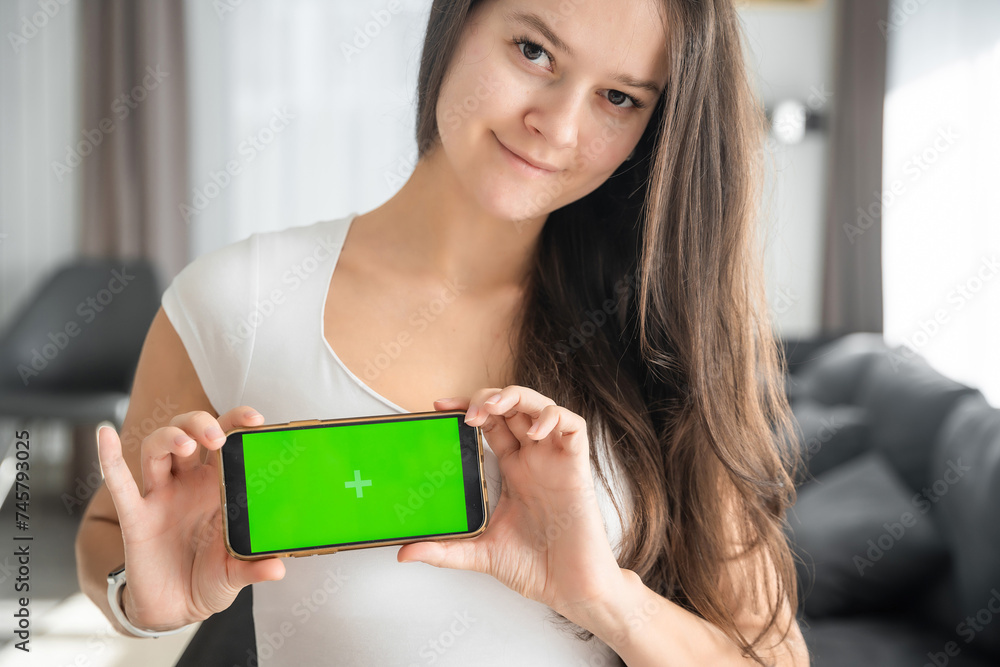 Young smiling woman shows smartphone with green screen in home living room closeup. Technology, gadget, connection. Advertising area, workspace mock up. Young smiling woman shows smartphone with green screen in home living room closeup. Technology, gadget, connection. Advertising area, workspace mock up.