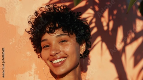 Smiling woman with curly hair and hoop earrings standing in front of a warm-toned wall with shadows from leaves.