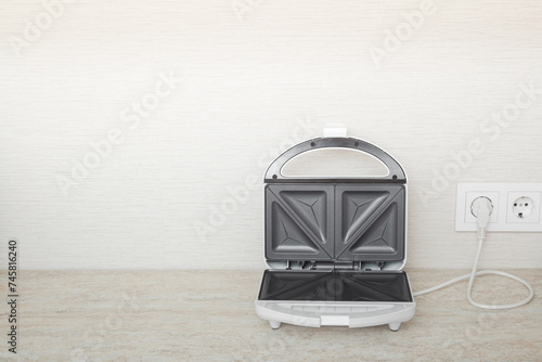 White opened sandwich maker with black nonstick surface with plug in wall outlet socket on table top at home kitchen. Closeup. Kitchen electric equipment. Empty place for text on wallpaper background.