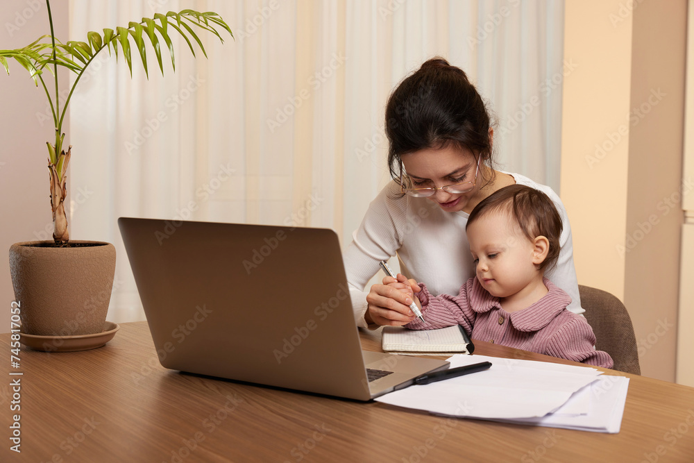 Cheerful pretty businesswoman working on laptop at home with her little child girl Cheerful pretty businesswoman working on laptop at home with her little child girl