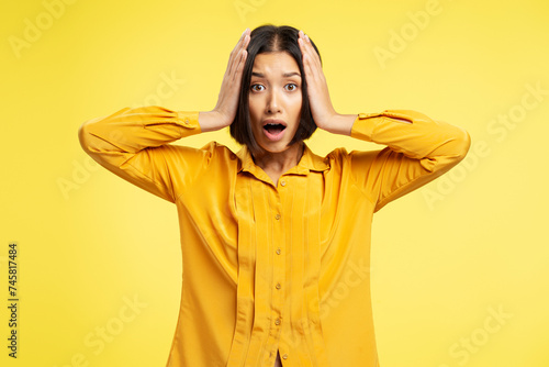 Portrait of serious, excited asian woman holding head with hands, looking at camera standing isolated on yellow background. Concept of advertisement, shopping, discounts © speed300