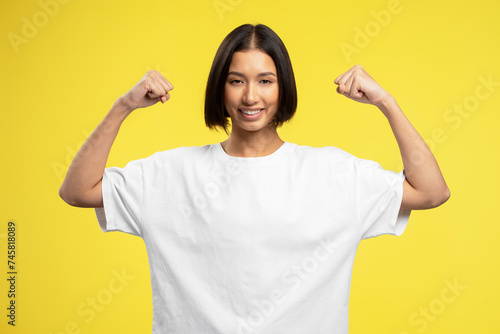 Smiling strong asian girl showing biceps, muscles standing isolated on yellow background