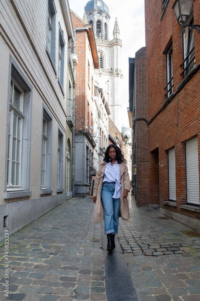 A young African woman is captured mid-stride walking down a narrow cobblestone street in an old European town. The historic charm of the town is exemplified by the traditional architecture and the