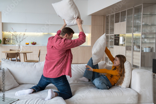 Happy joyful couple fighting with pillows on sofa in modern apartment, laughing, having fun, joy happiness, positive emotions. Emotional moments in relations. Spending active weekends together at home photo