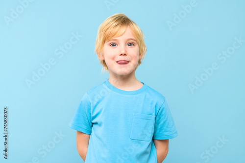 Funny young boy showing tongue and looking at the camera over pastel blue background. Cute preschooler grimacing at camera sticking out his tongue.