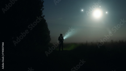 A mysterious figure with torch looking at glowing UFO lights in the sky. On a spooky misty night in the countryside. photo