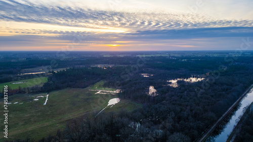 Fototapeta Naklejka Na Ścianę i Meble -  This image provides a breathtaking aerial view of a tranquil landscape at sunset. The sun dips towards the horizon, its rays filtering through the clouds, casting a warm glow over the land. Water
