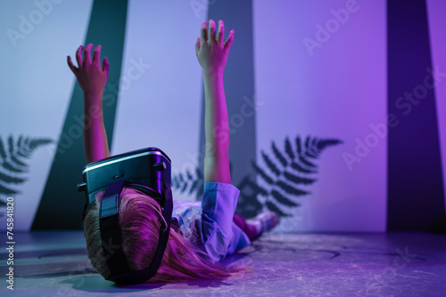 Child girl with virtual reality glasses laying on the VR room floor, lit by fluorescent light effects treetop grow simulation. Science and children concepts.
